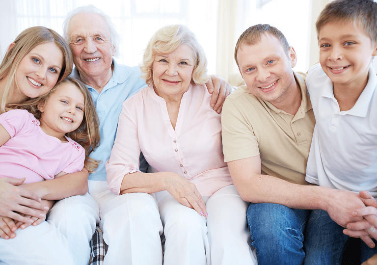 Drei Generationen einer Familie sitzen eng beieinander auf einem Sofa und lächeln in die Kamera: Großeltern, Eltern und zwei Kinder.
